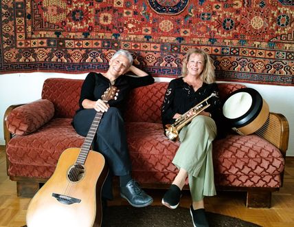 Zwei Frauen sitzen entspannt auf einem roten Sofa. Eine hält eine Gitarre, die andere eine Trompete und eine Trommel. Im Hintergrund hängt ein bunter Teppich.