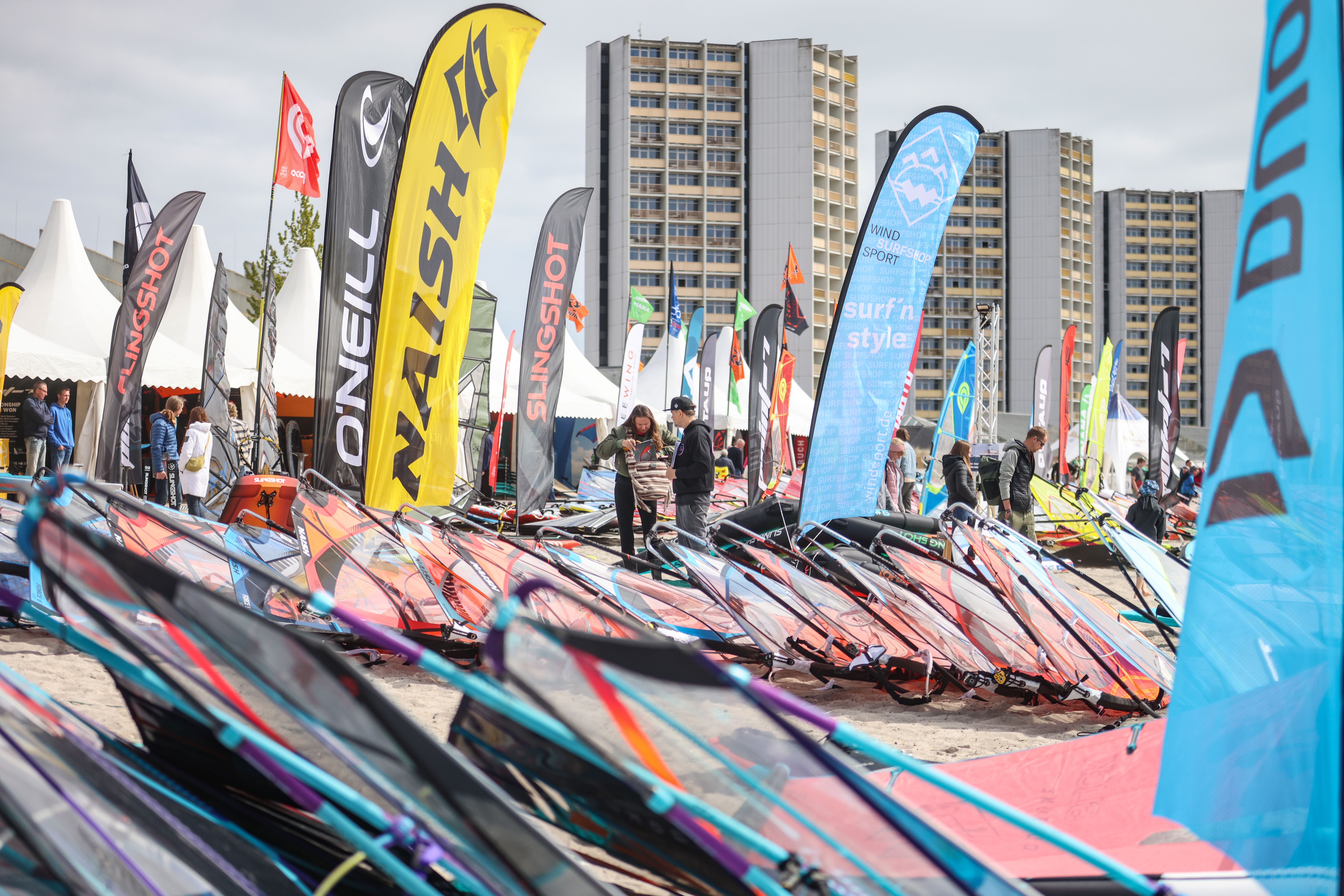 Bunte Surfsegel liegen am Strand. Im Hintergrund wehen bunte Fahnen