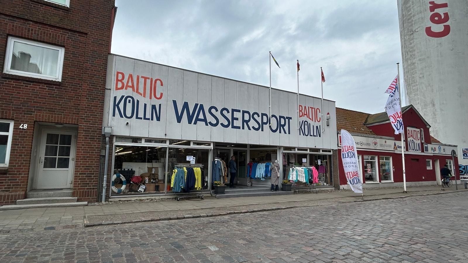 Ein Wassersportgeschäft namens 'Baltic Kölln' mit Schaufenstern voller Kleidung und Ausrüstung. Das Gebäude hat eine große Beschriftung und Flaggen.