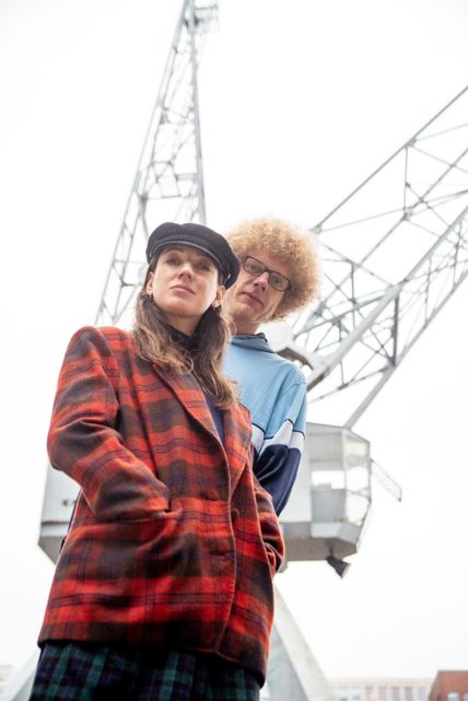 Eine Frau mit rot karierter Jacke und ein mann mit hellblauem Pullover, stehen versetzt hintereinander. Im Hintergrund ist ein Kran zu sehen.