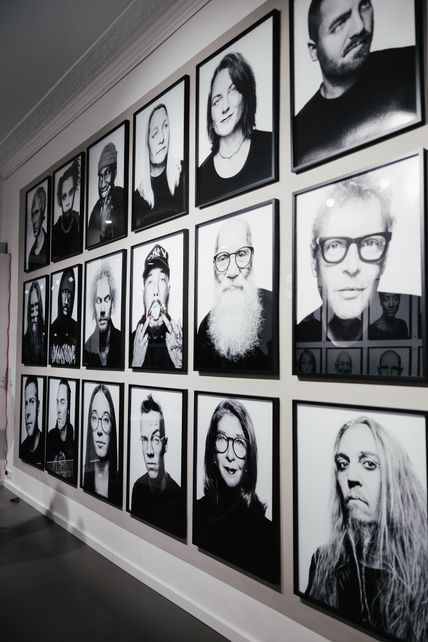 Eine Galerie mit schwarz-weißen Porträtfotos von verschiedenen Personen, die in einem Raster an der Wand hängen. Die Bilder sind gerahmt und zeigen diverse Gesichtsausdrücke.