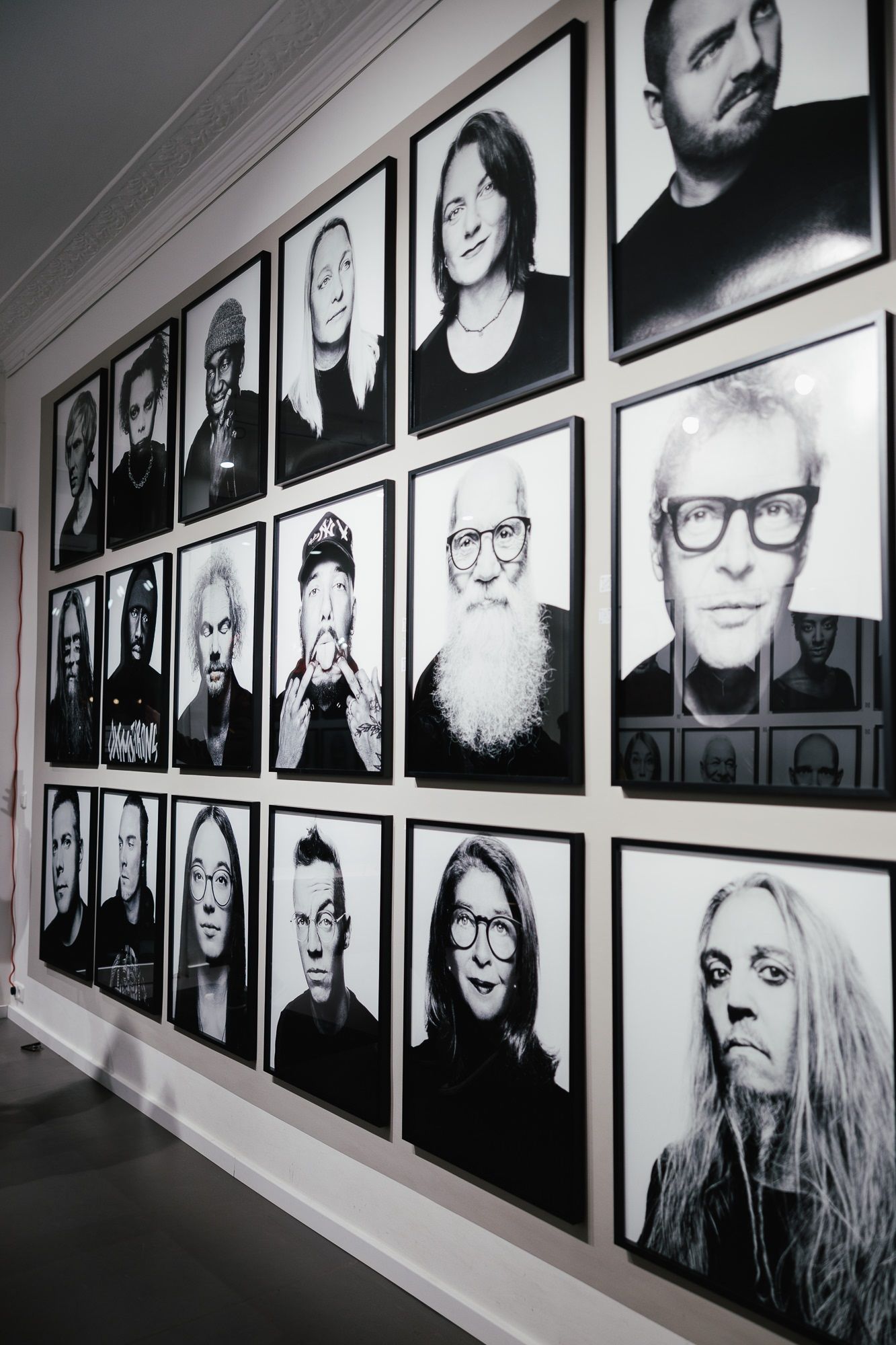 Eine Galerie mit schwarz-weißen Porträtfotos von verschiedenen Personen, die in einem Raster an der Wand hängen. Die Bilder sind gerahmt und zeigen diverse Gesichtsausdrücke.