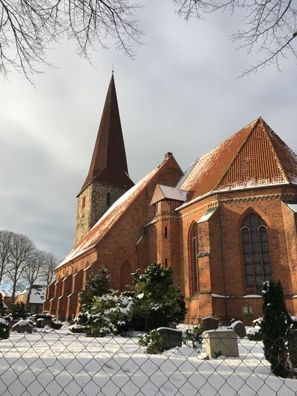 Die St. Johannis-Kirche in Petersdorf ist im Winter von Schnee bedeckt. Der rote Backsteinbau mit hohem Turm steht unter einem bewölkten Himmel.