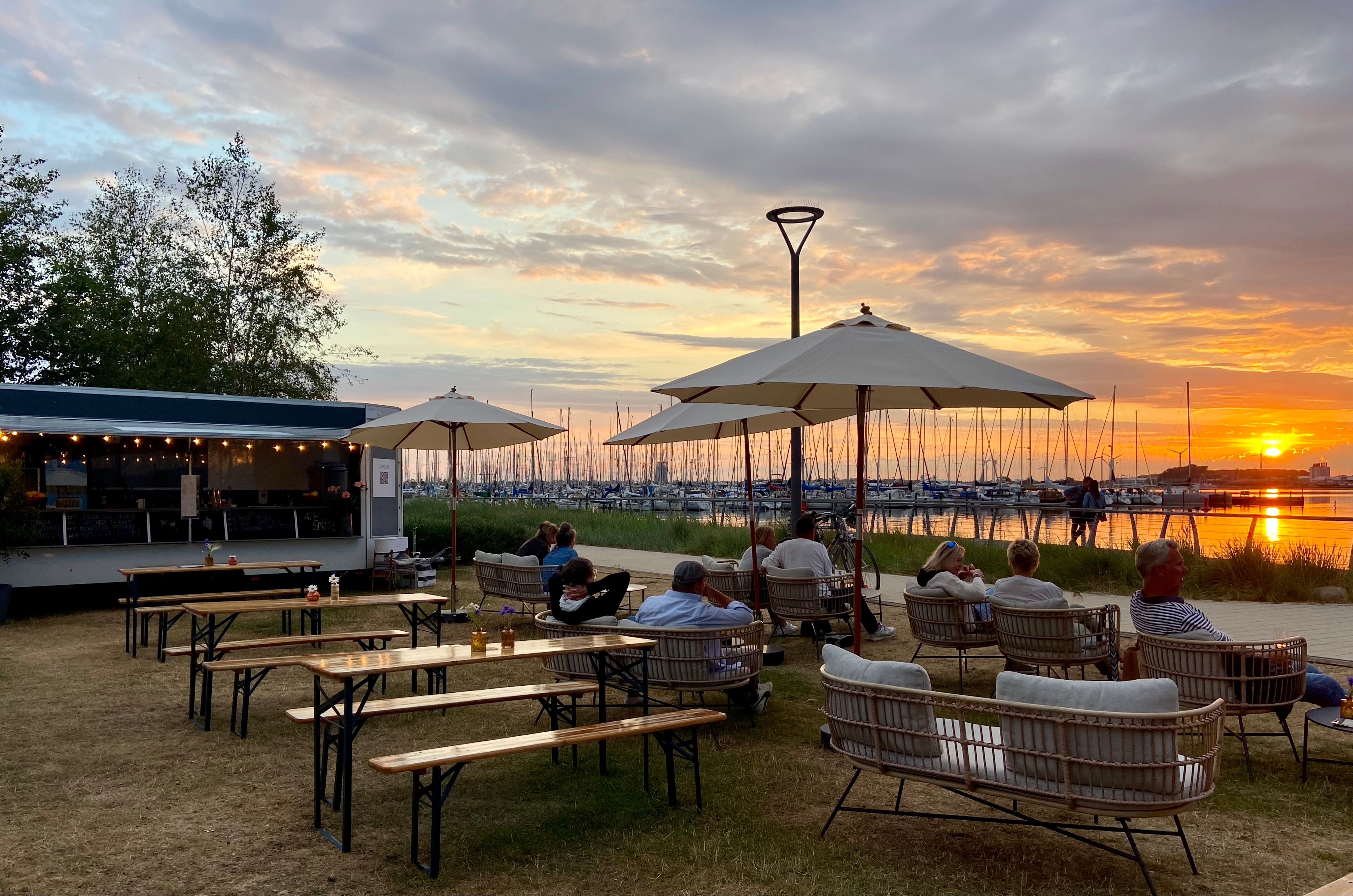 Gemütliche Sitzgelegenheiten und Sonnenschirme am Hafen bei Sonnenuntergang. Menschen genießen die Aussicht auf Boote und das Wasser.