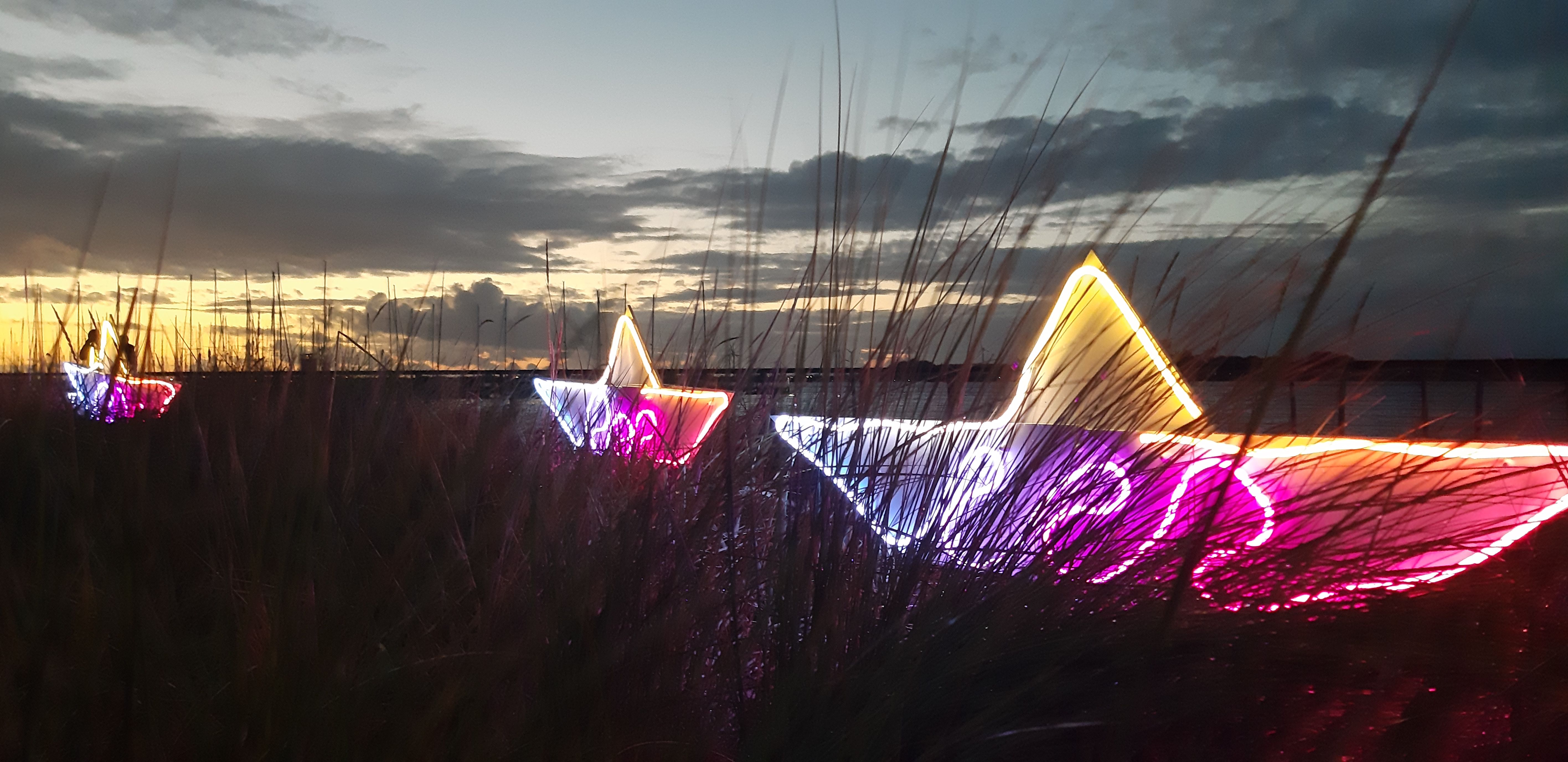 Leuchtende Papierboote in verschiedenen Farben stehen in einer Wiese bei Sonnenuntergang. Der Himmel ist bewölkt und die Boote strahlen hell.