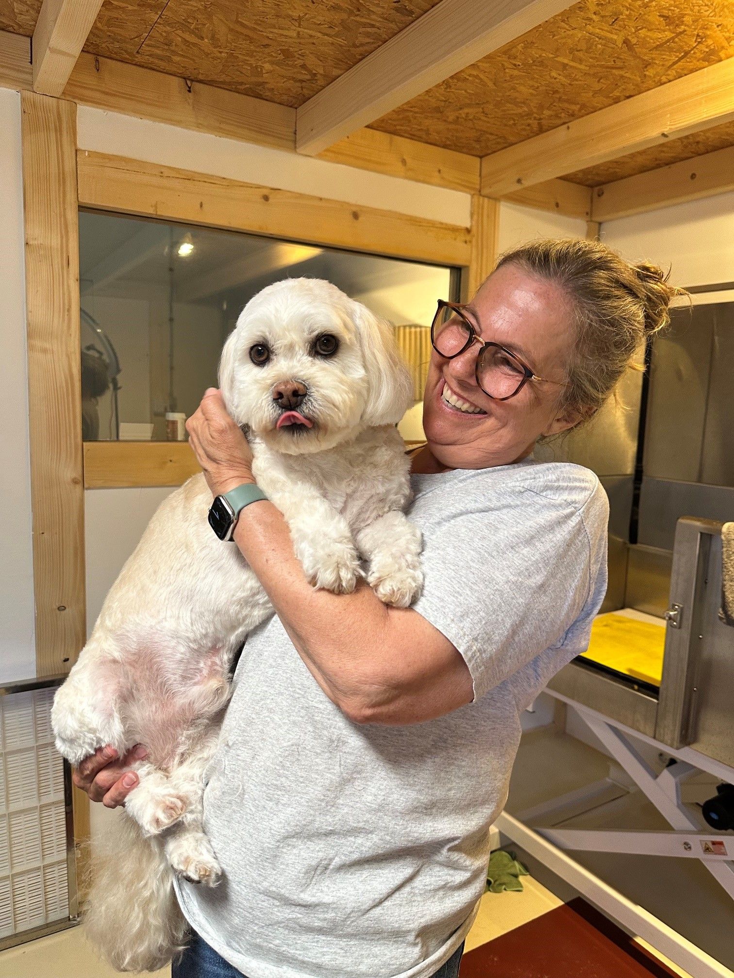 Eine Frau in einem Hundesalon hält einen frisch geschnittenen weißen Hund und lächelt. Der Hund streckt die Zunge heraus.