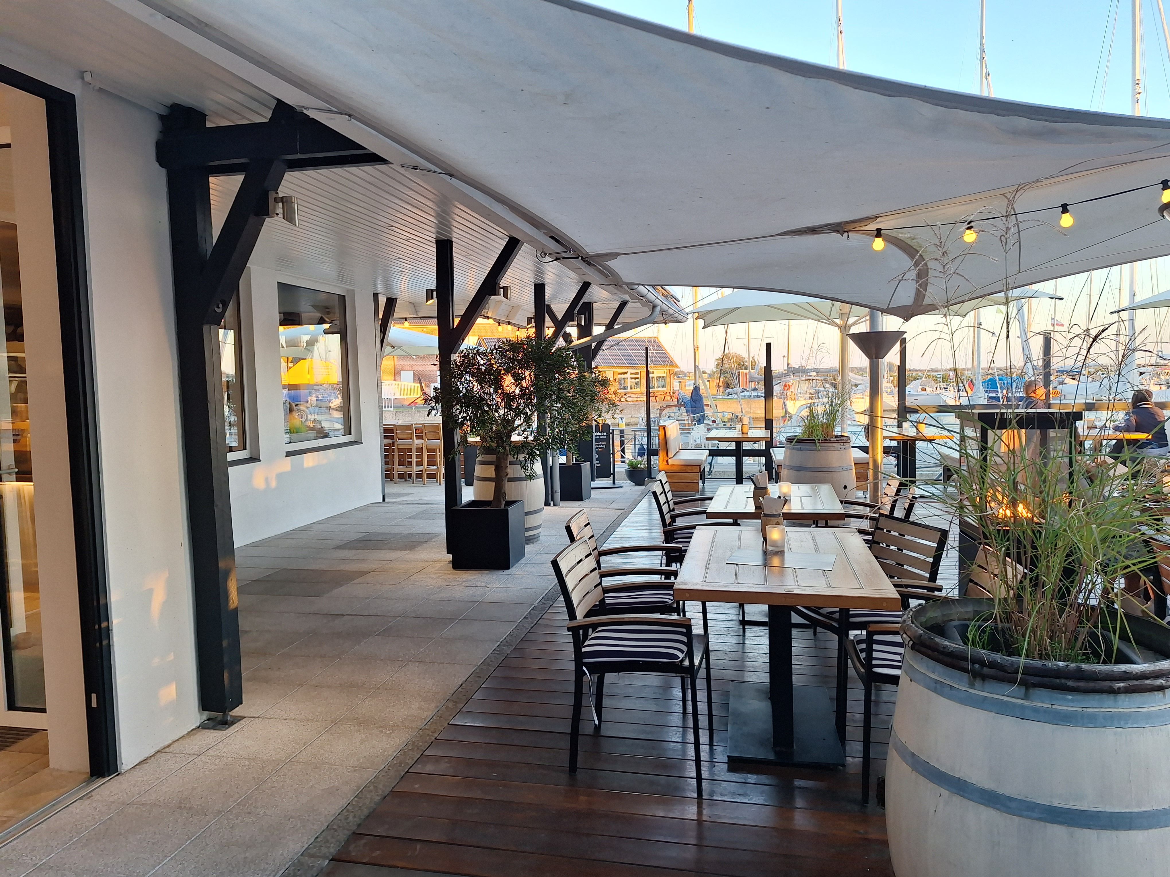 Gemütlicher Außenbereich eines Restaurants am Hafen von Lemkenhafen mit Holztischen, Stühlen und Segeltuchüberdachung. Im Hintergrund sind Segelboote zu sehen.