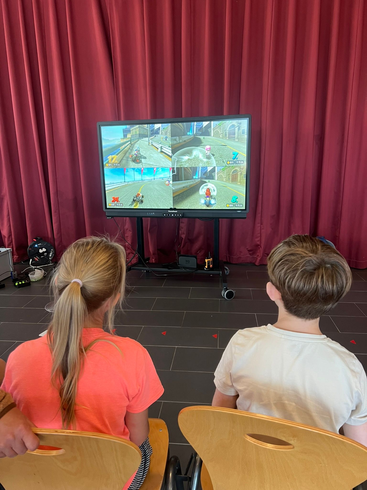 Zwei Kinder spielen ein Videospiel auf einem großen Bildschirm. Sie sitzen auf Holzstühlen vor einem roten Vorhang.