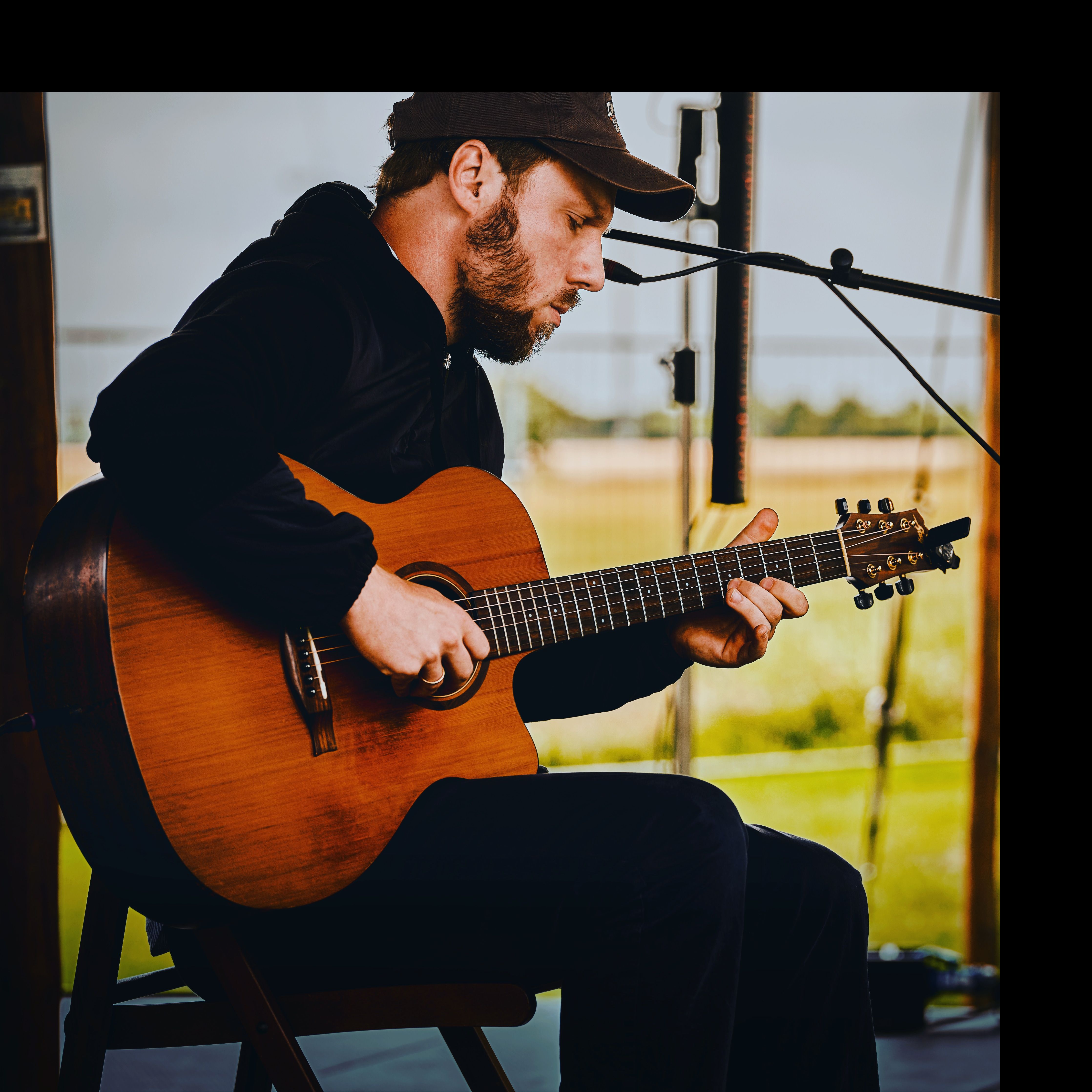 Ein Musiker spielt konzentriert Akustikgitarre auf einer Bühne im Freien. Er trägt eine Mütze und sitzt auf einem Stuhl.