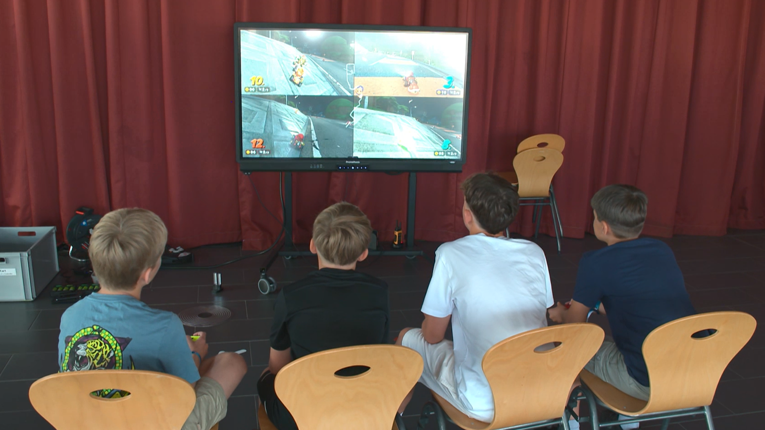 Vier Kinder spielen Mario Kart auf einem großen Bildschirm. Sie sitzen auf Stühlen in einem Raum mit roten Vorhängen.