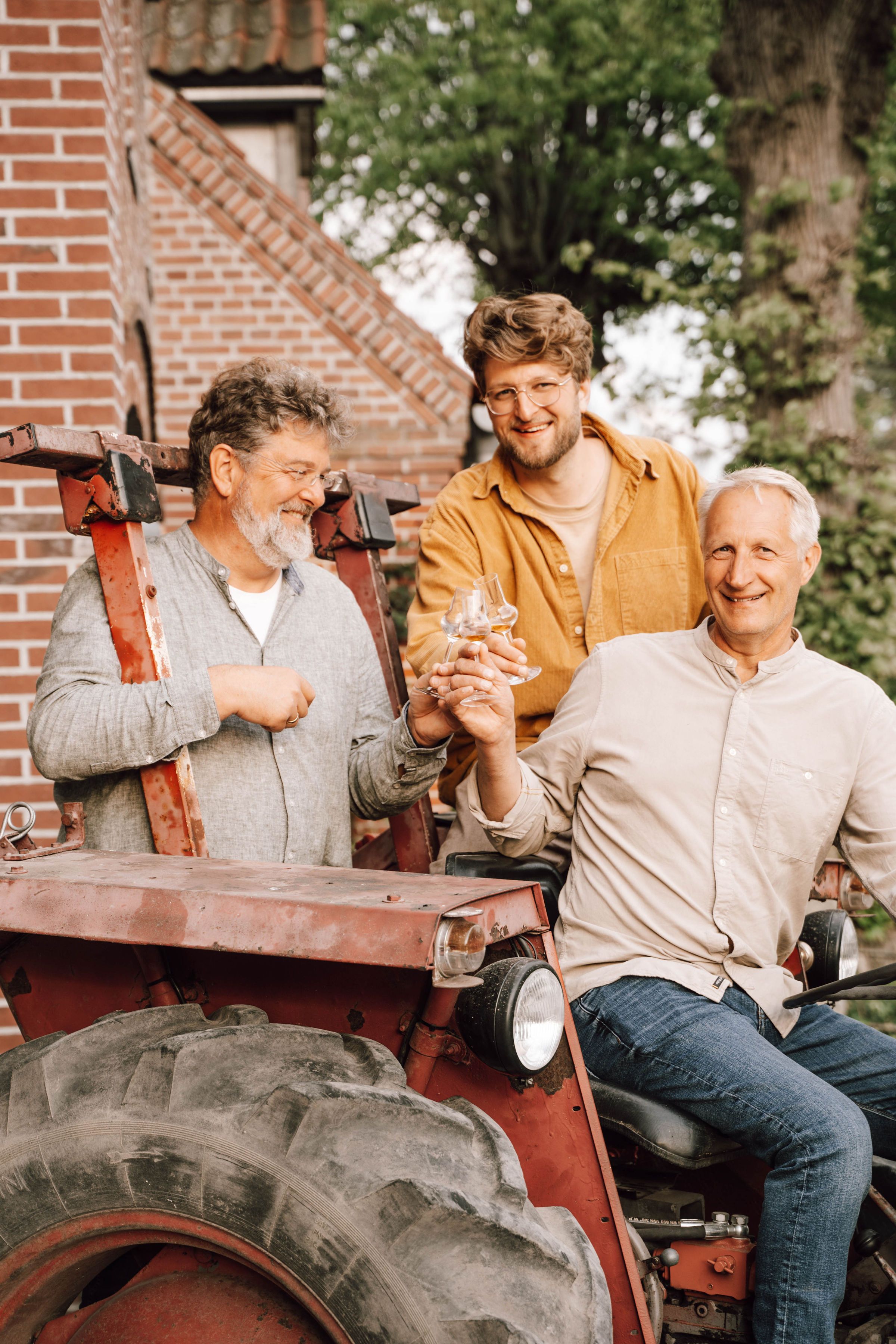 Drei Männer sitzen auf einem alten Traktor vor einem Backsteingebäude und stoßen mit Gläsern an. Sie wirken fröhlich und entspannt.