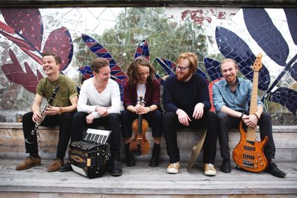 Fünf Musiker sitzen lachend auf einer Bank vor einem bunten Mosaik. Sie halten verschiedene Instrumente wie Klarinette, Akkordeon, Geige, Becken und Bassgitarre.
