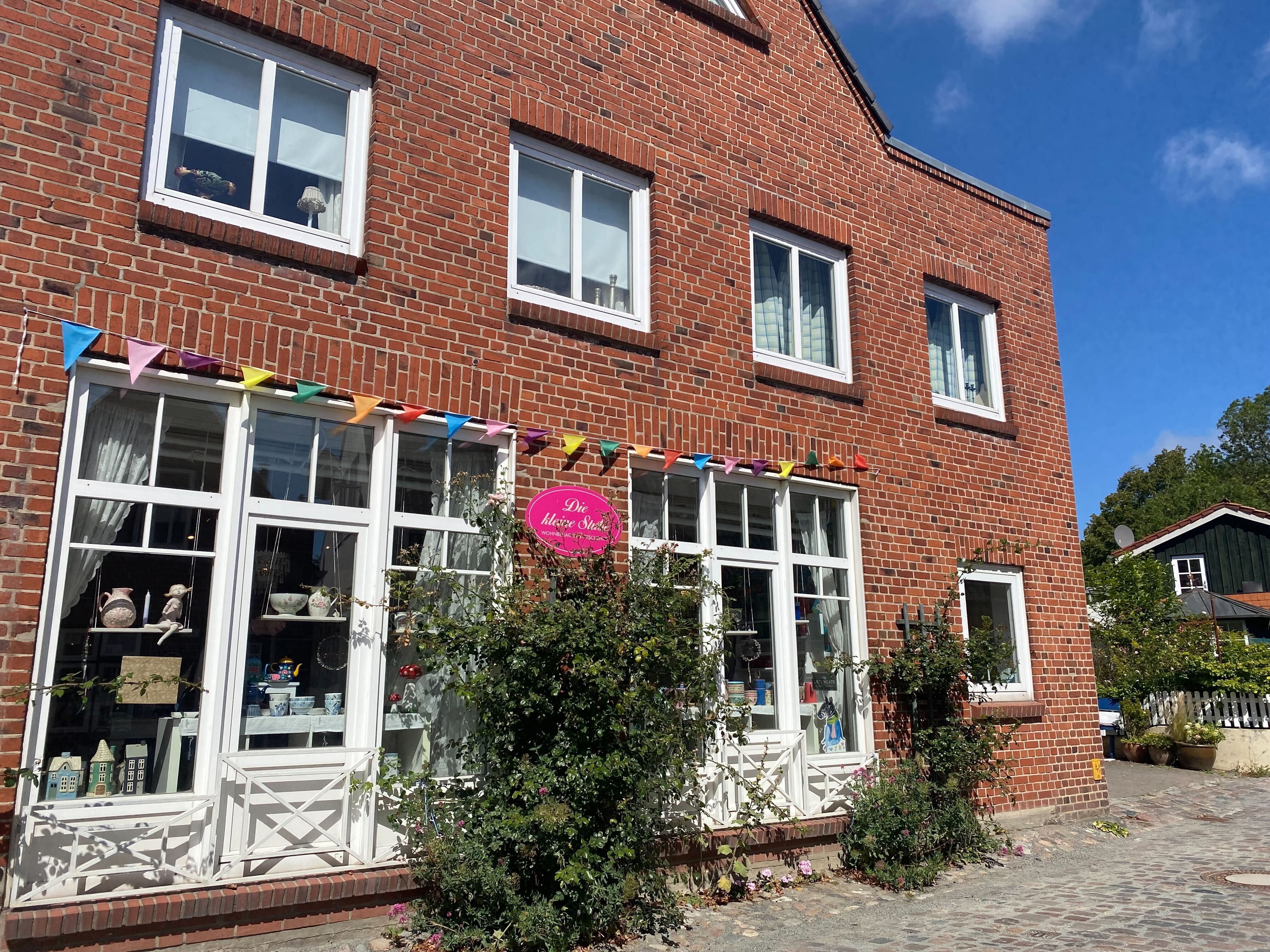 Backsteingebäude mit Schaufenster, bunter Wimpelkette und Pflanzen. Ein Schild mit der Aufschrift 'Die kleine Stube' ist sichtbar.