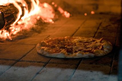 Eine Pizza wird in einem traditionellen Holzofen gebacken. Der Ofen ist mit brennendem Holz beheizt, das eine warme, einladende Atmosphäre schafft.