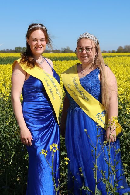 Zwei Frauen in blauen Kleidern und gelben Schärpen stehen in einem blühenden Rapsfeld unter blauem Himmel.