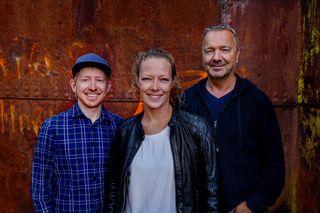 Drei Personen stehen lächelnd vor einer rostigen Metallwand. Eine Frau in der Mitte, flankiert von zwei Männern. Alle wirken entspannt und freundlich.