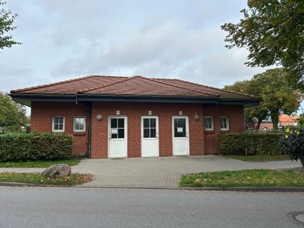 Kleines Backsteingebäude mit roten Dachziegeln, drei weißen Türen und kleinen Fenstern, umgeben von Bäumen und Sträuchern.