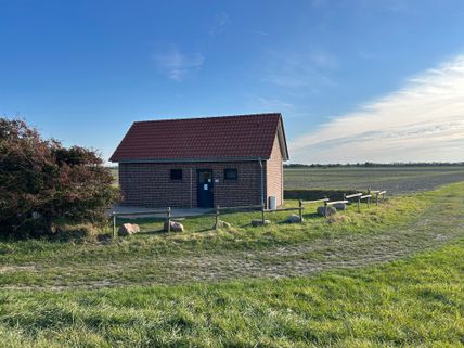 Ein kleines Backsteingebäude mit rotem Ziegeldach steht auf einem Feld unter blauem Himmel. Ein Weg führt daran vorbei, umgeben von Gras und Büschen.