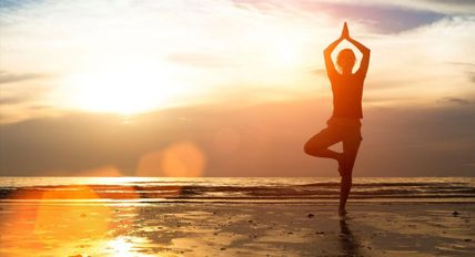 Eine Person steht in einer Yoga-Pose am Strand, während die Sonne im Hintergrund untergeht. Die Szene strahlt Ruhe und Gelassenheit aus.