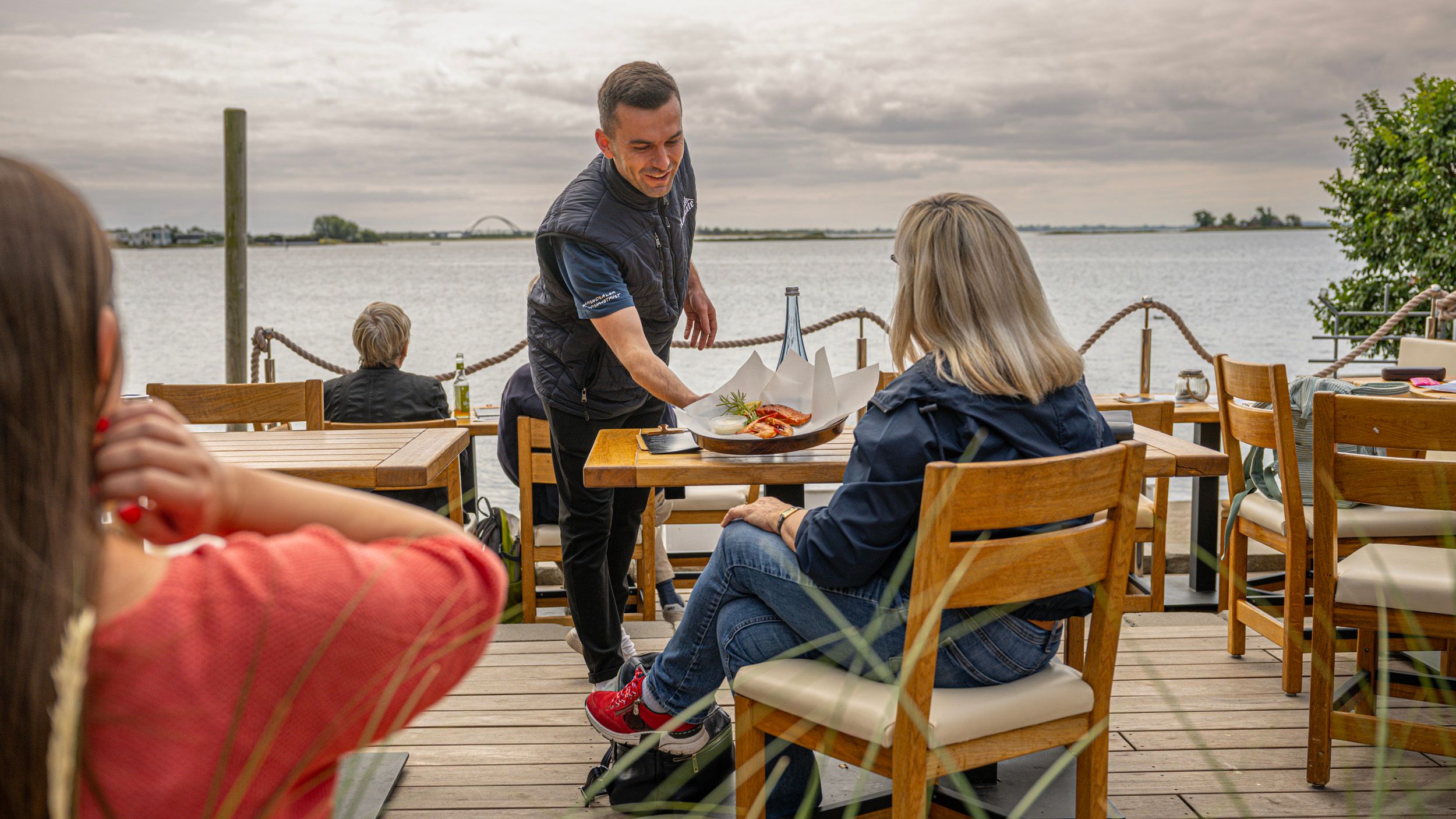 Ein Kellner serviert einem Gast auf einer Terrasse mit Blick auf das Wasser ein Gericht. Im Hintergrund ist eine Brücke zu sehen.