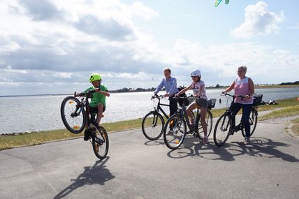 Eine Familie radelt fröhlich auf einem Deich entlang, während ein Kind einen Wheelie macht. Im Hintergrund ist das Meer zu sehen.