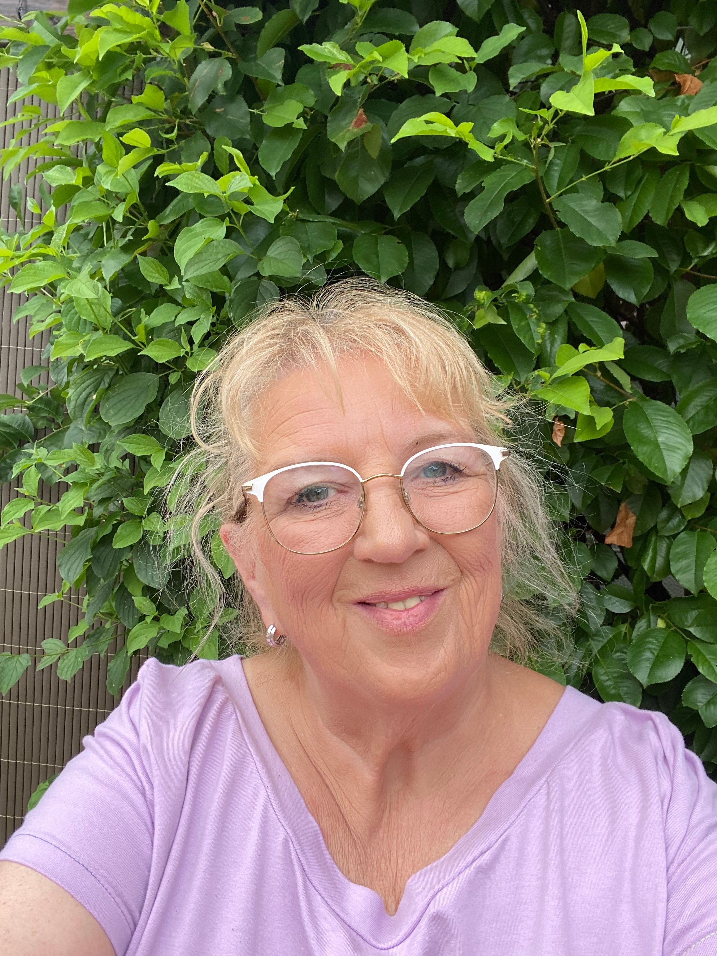 Eine lächelnde Frau mit Brille und lila Oberteil steht vor einer dichten, grünen Hecke.