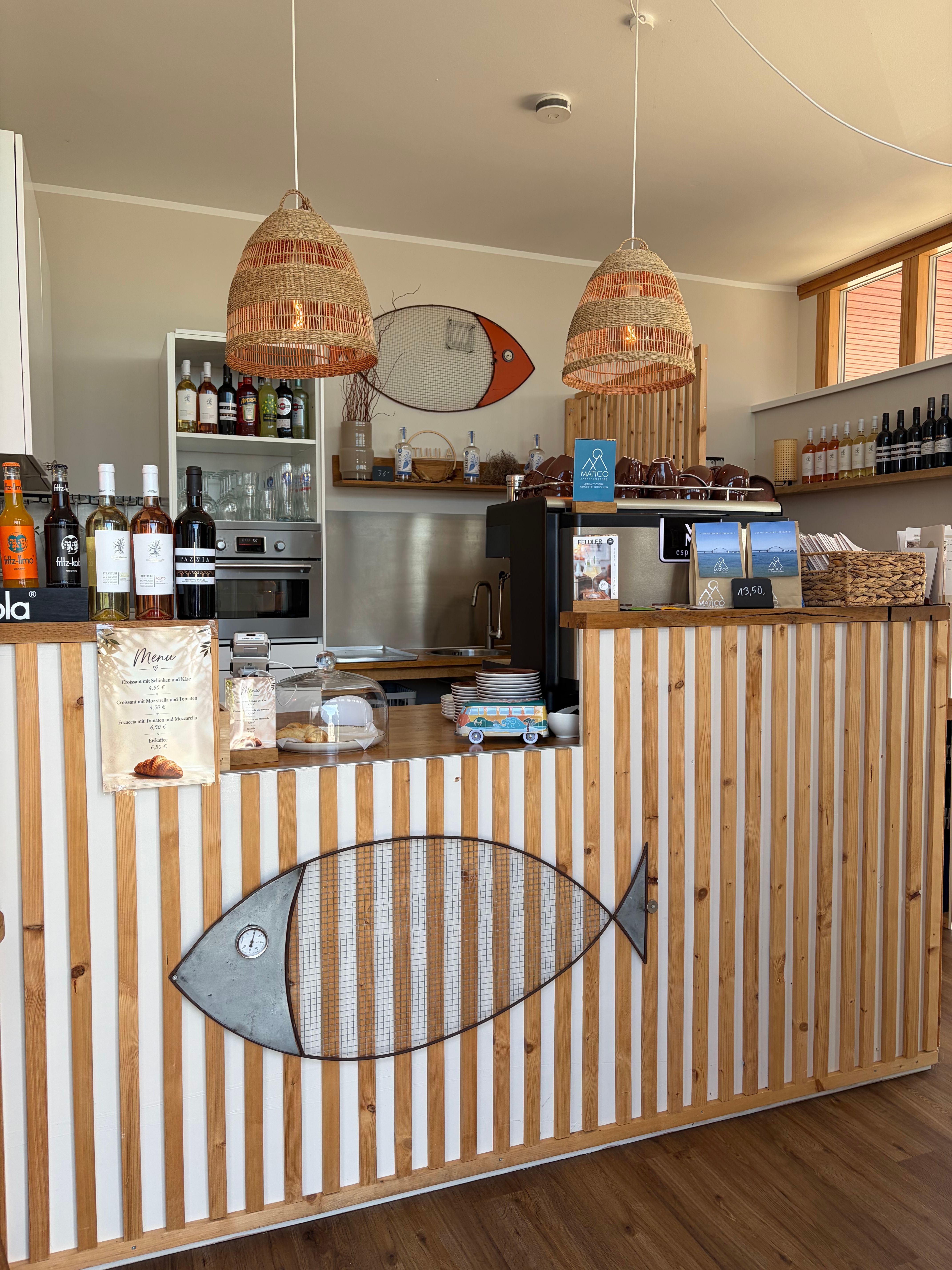 Ein gemütliches Café mit Holztresen, dekoriert mit Fischmotiven. Hängelampen aus Korbgeflecht und eine Auswahl an Getränken sind sichtbar.