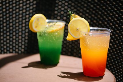 Zwei erfrischende Getränke in Plastikbechern, eines grün und eines orange, mit Zitronenscheiben und Rosmarin garniert, stehen auf einem Tisch.