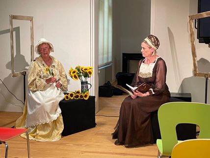 Zwei Frauen in historischen Kostümen sitzen auf einer Bühne. Eine hält Sonnenblumen, die andere ein Buch. Im Hintergrund sind leere Bilderrahmen.