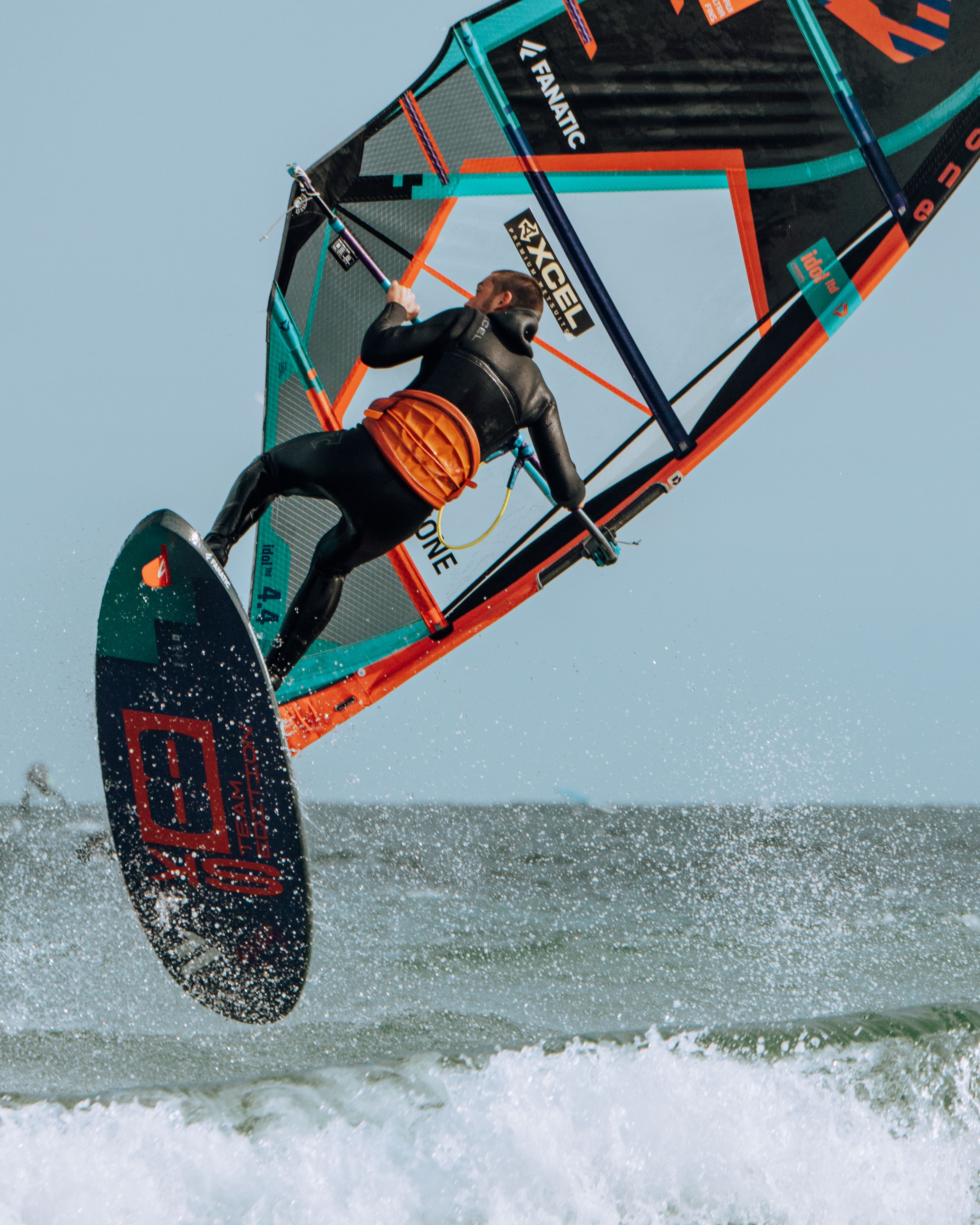 Ein Windsurfer in Aktion, springt über eine Welle. Er trägt einen Neoprenanzug und hält das Segel fest. Der Himmel ist klar.