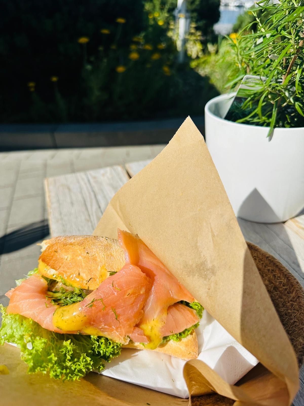 Ein frisches Lachsbrötchen mit Salat und Sauce, serviert auf einem Holztisch im Freien. Daneben steht ein Topf mit Kräutern.