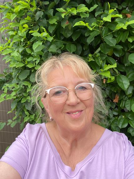 Eine lächelnde Frau mit Brille und lila Oberteil steht vor einer dichten, grünen Hecke.