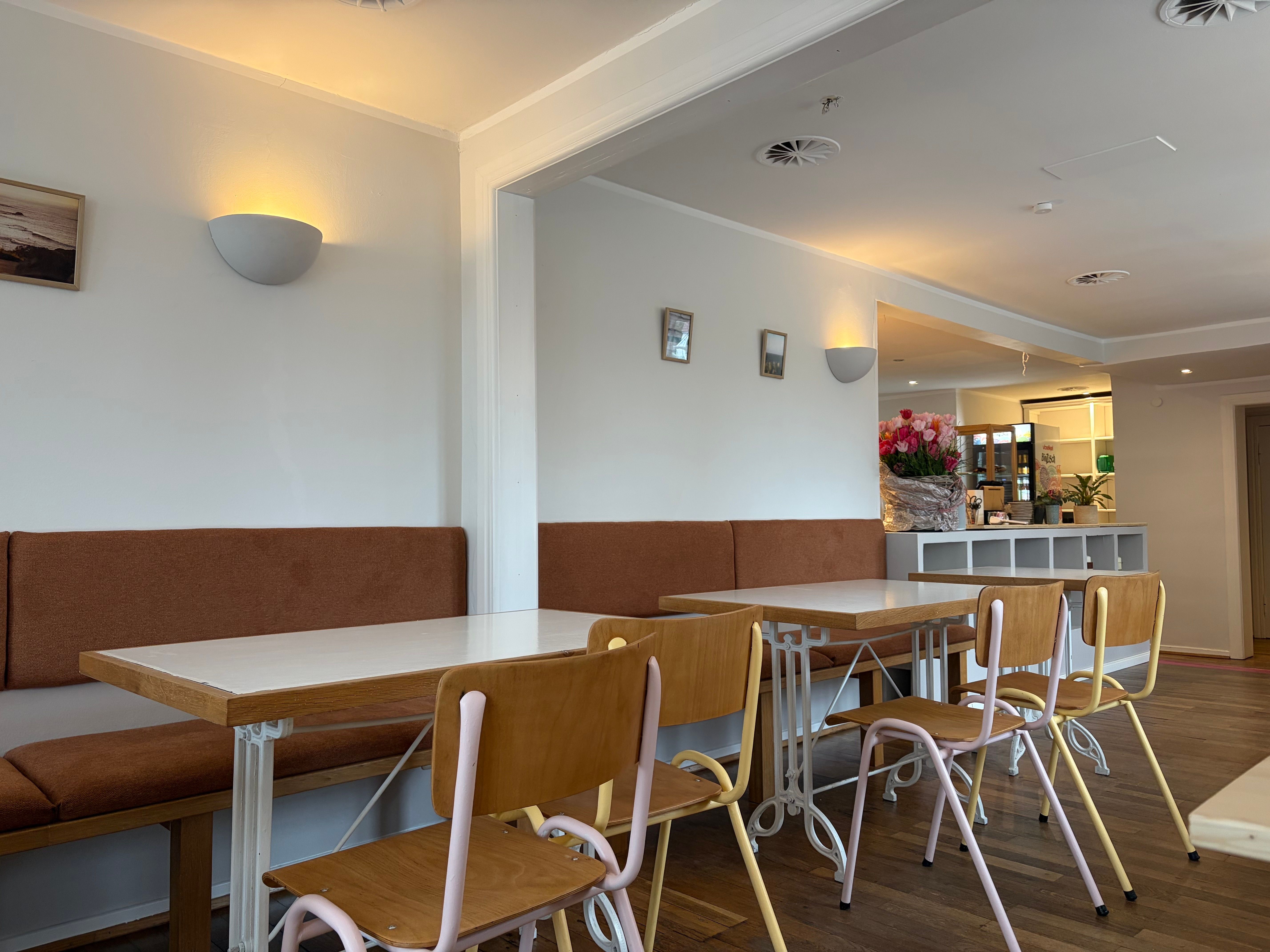 Ein gemütliches Café mit Holztischen, Stühlen und braunen Sitzbänken. An der Theke stehen Blumen, die Atmosphäre ist einladend und hell.